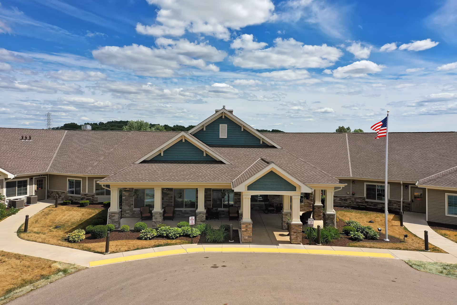 Front exterior view of Beloit Senior Living - The Gardens building with a covered entrance, stone pillars, landscaped bushes, an American flag on a flagpole, and a partly cloudy sky above.