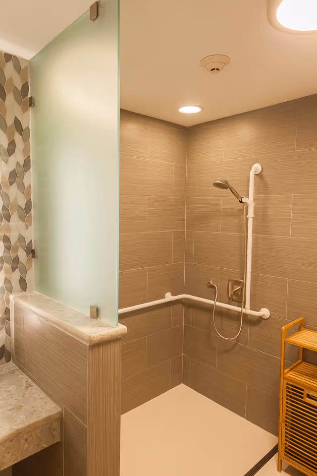 A modern shower area with beige tiled walls and a handheld showerhead mounted on a vertical grab bar. There is a horizontal grab bar along the wall for support. The shower area has a frosted glass partition separating it from a countertop with a marble surface. A wooden shelving unit is placed next to the shower. The ceiling has recessed lighting and a smoke detector.