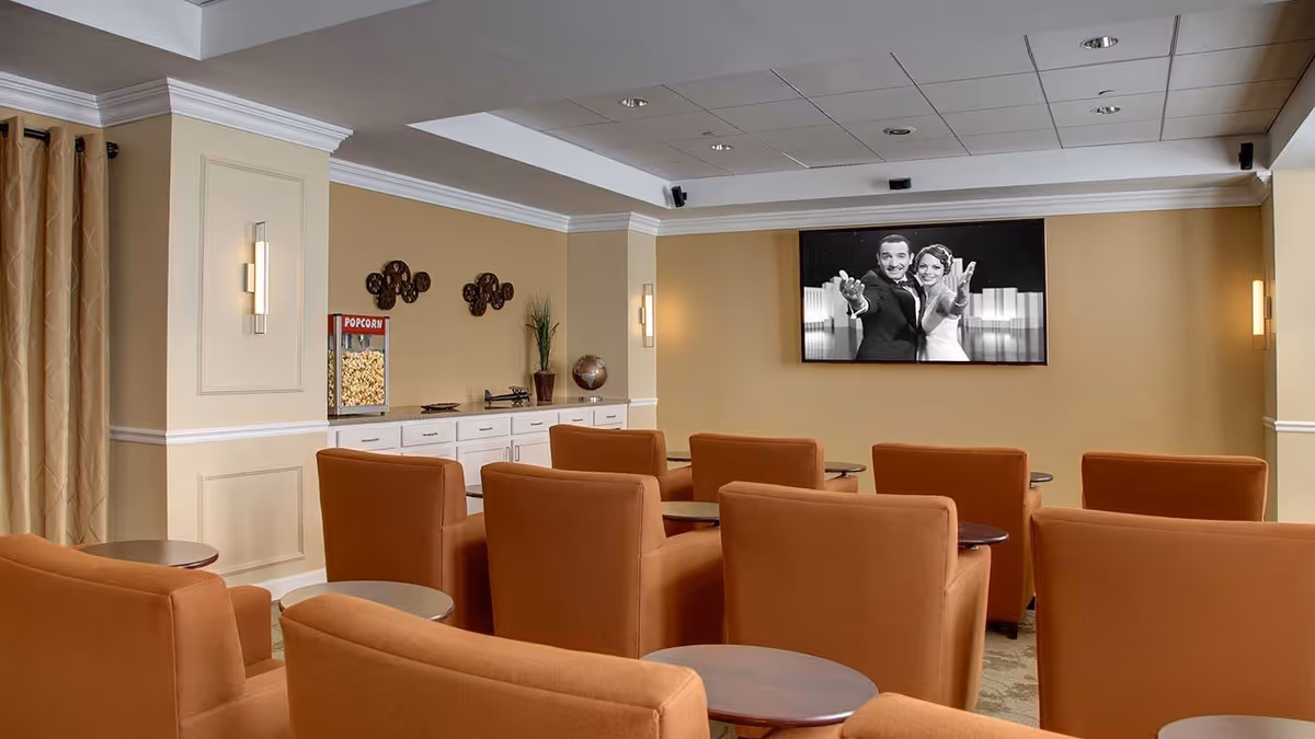 A cozy movie theater room with multiple rows of orange upholstered chairs facing a large screen displaying a black and white image of a man and woman dancing. There is a popcorn machine on a white cabinet against the beige wall, along with decorative items and wall sconces providing soft lighting.