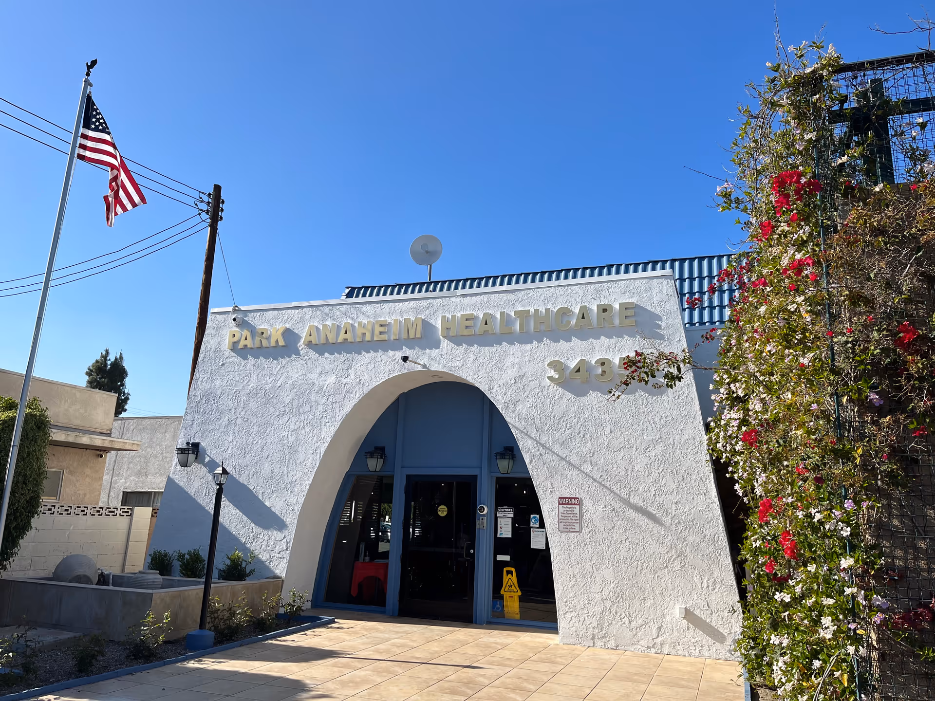 Exterior view of Park Anaheim Healthcare building with a white stucco facade and a large archway entrance. An American flag is flying on a flagpole to the left, and there are flowering plants climbing a trellis on the right side. The sky is clear and blue.