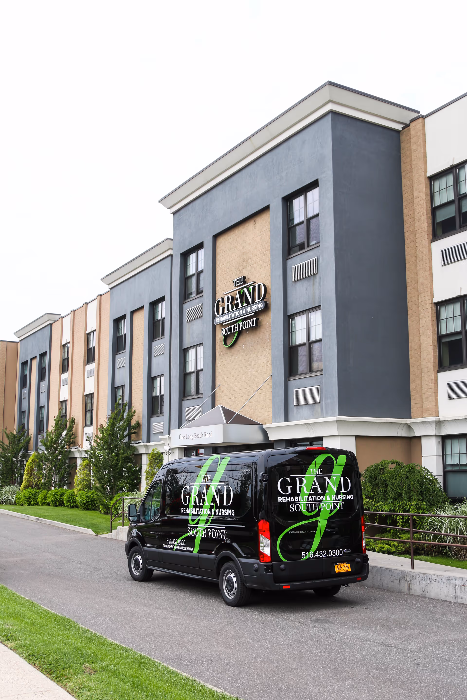 Exterior view of The Grand Rehabilitation and Nursing at South Point building with a black shuttle van parked in front. The building is multi-story with beige and gray walls and several windows. The van displays the facility's name and contact information.
