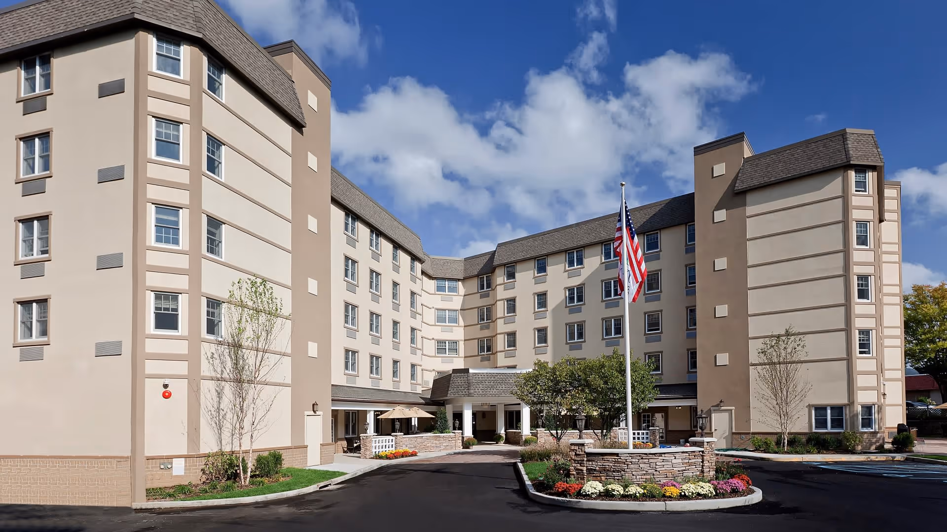 Exterior view of a multi-story senior living facility building with beige walls and multiple windows under a partly cloudy blue sky. There is a circular driveway with a landscaped island in the center featuring an American flag and colorful flowers. Small trees and shrubs are planted around the building.