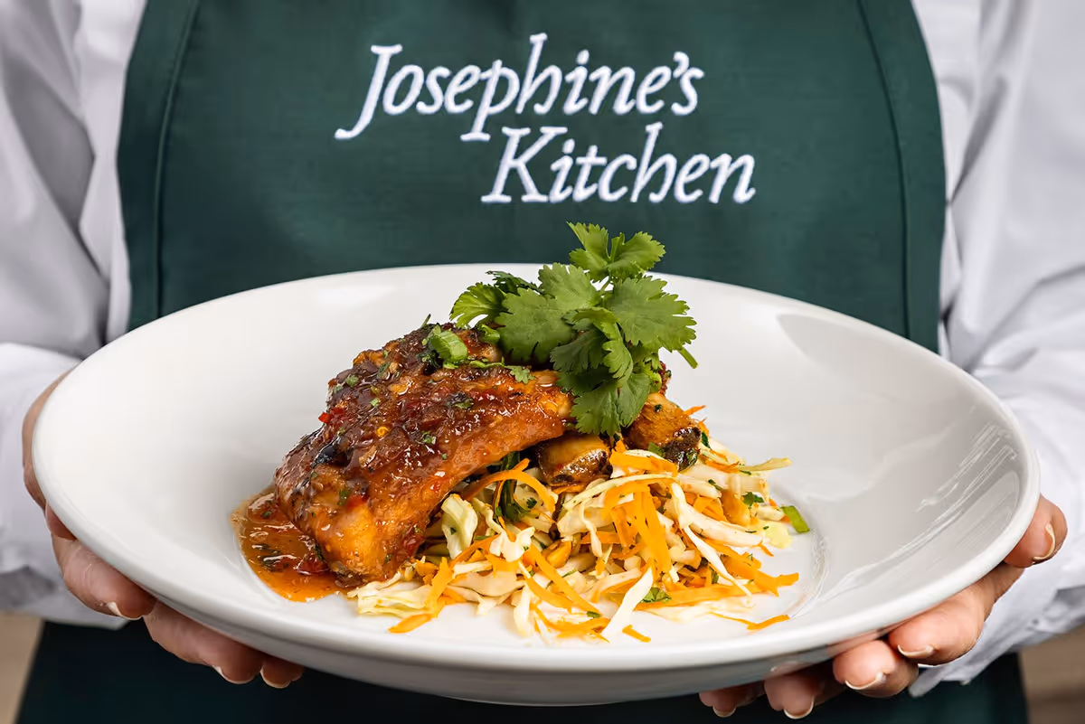 A person wearing a green apron with the text 'Josephine's Kitchen' holds a white plate with a serving of cooked fish topped with sauce and garnished with fresh cilantro, placed on a bed of shredded vegetables including cabbage and carrots.