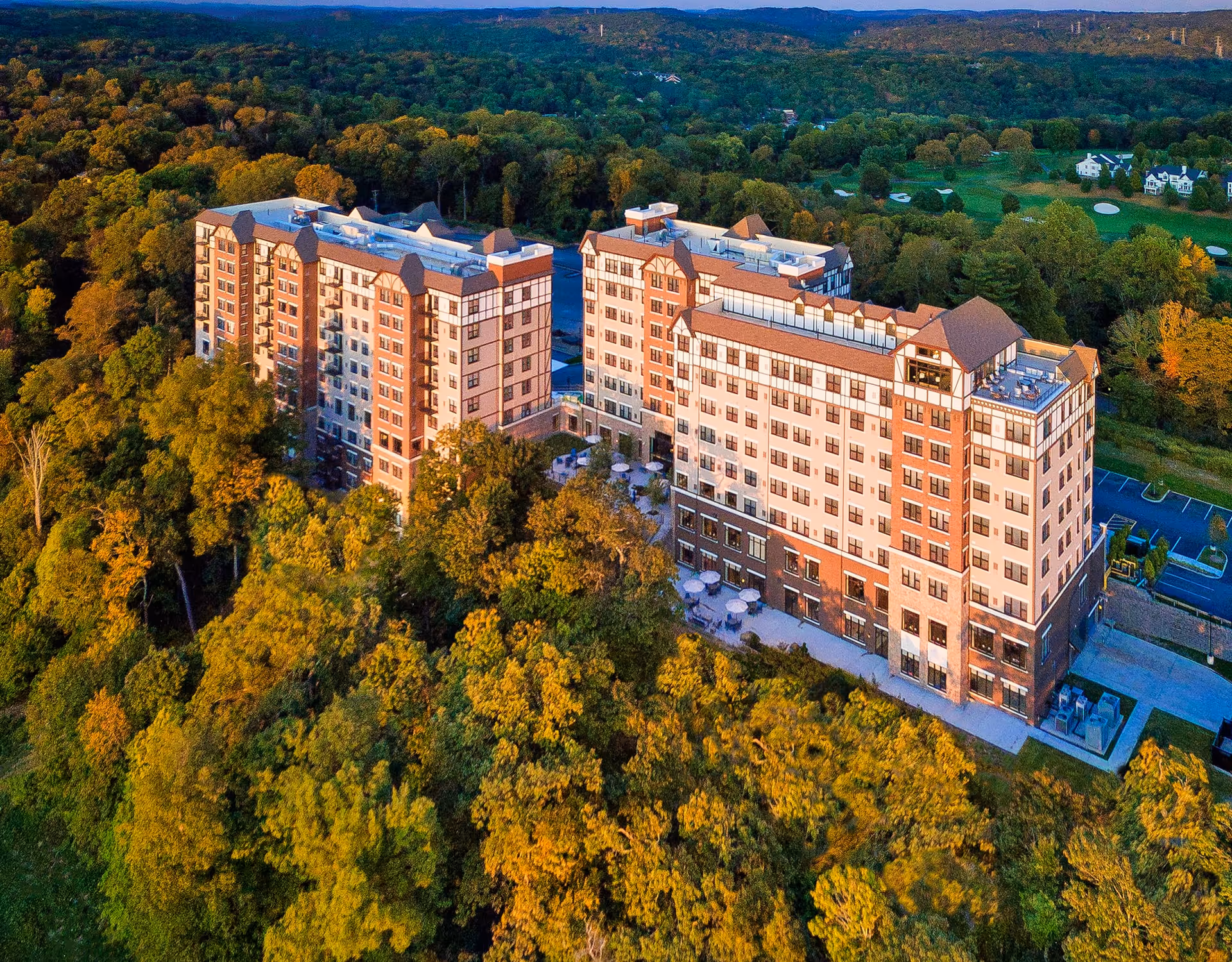 Aerial view of a large senior living facility named The Club at Briarcliff Manor surrounded by dense green and autumn-colored trees, with a golf course visible in the background and a parking lot adjacent to the building.