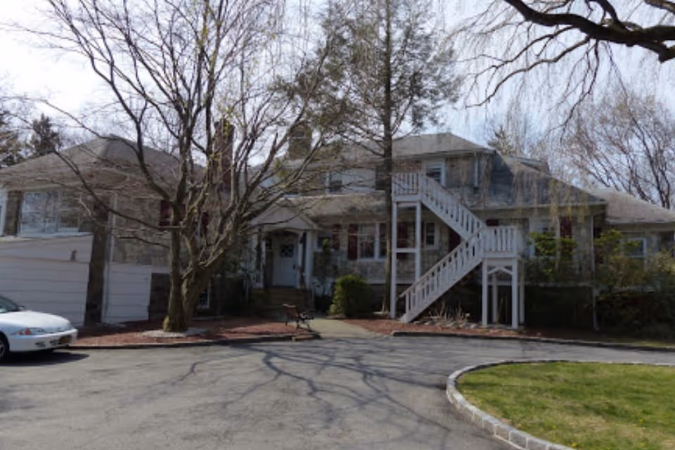 Exterior view of a two-story residential building with stone walls, multiple windows, and a white exterior staircase leading to the second floor. There is a large leafless tree in front, a white car parked on the left, and a curved driveway with some green grass on the right.