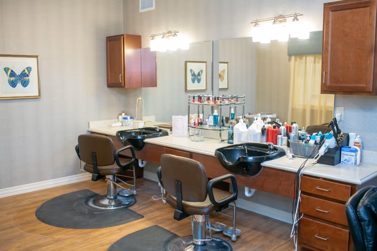 Interior view of a senior living facility's hair salon area with two brown salon chairs in front of a counter with two black hair washing sinks. The counter holds various hair care products, nail polish, and styling tools. There are two wall-mounted cabinets and a large mirror above the counter. A framed butterfly picture hangs on the wall.