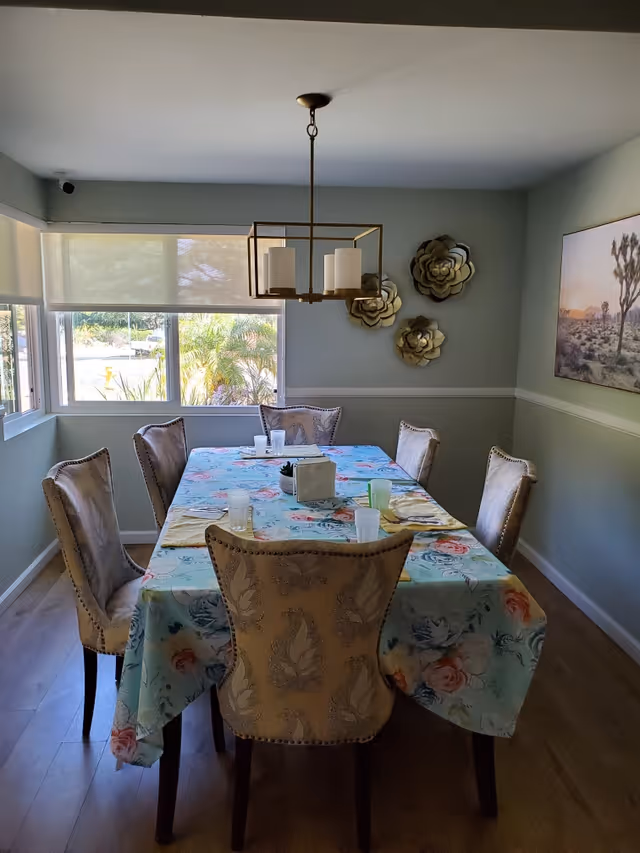 A dining room with a rectangular table covered with a floral tablecloth. Six upholstered chairs surround the table. On the table are several cups, napkins, and a small centerpiece. The room has light-colored walls with decorative wall art including three metallic flower sculptures and a framed picture of a desert landscape. A modern chandelier with four candle-style lights hangs above the table. Large windows with roller shades allow natural light into the room.