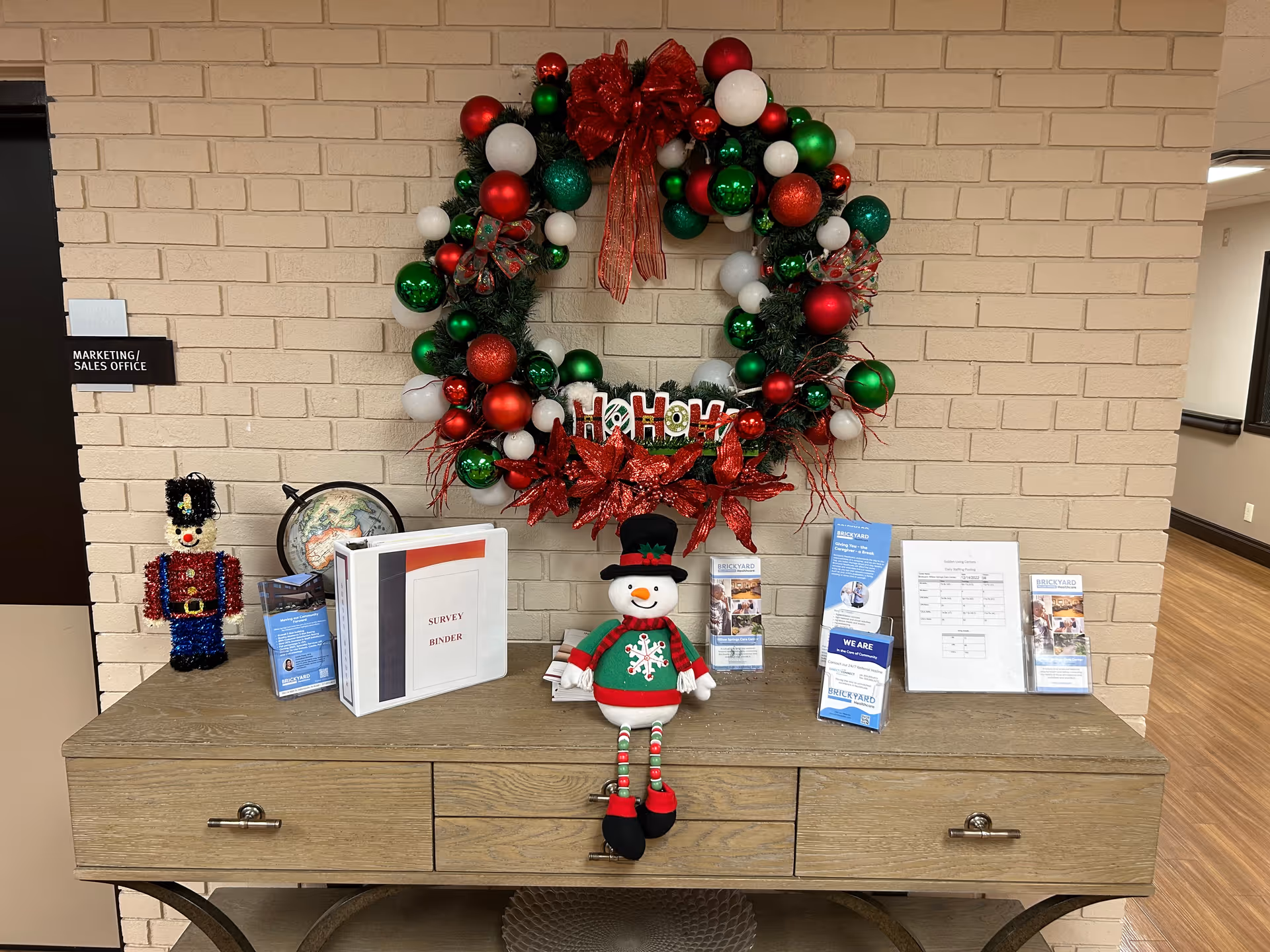 A wooden console table against a beige brick wall decorated for Christmas with a large wreath adorned with red, green, and white ornaments and a red bow. On the table are a festive snowman doll, a toy soldier figure, a globe, a survey binder, brochures, and a framed document. A sign on the left wall reads 'Marketing / Sales Office'.