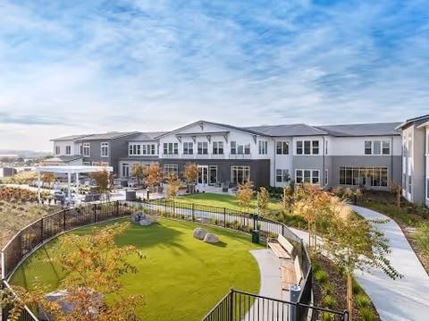 Exterior view of a modern assisted living and memory care facility with a two-story building surrounding a landscaped outdoor area featuring a green lawn, trees, benches, and a walking path under a partly cloudy sky.