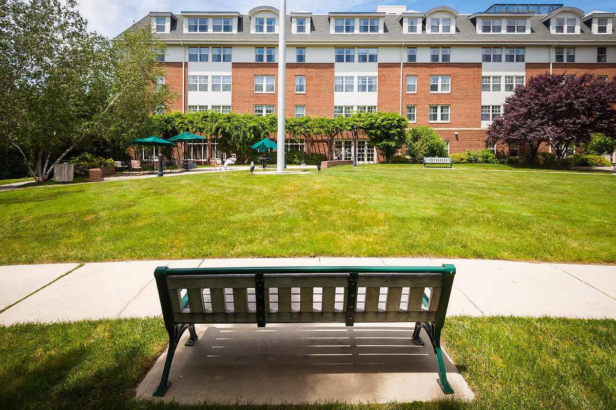 View of a green bench facing a large grassy lawn with a flagpole in the center, surrounded by trees and patio umbrellas. In the background, there is a multi-story brick and white building with many windows, part of Providence House Assisted Living.