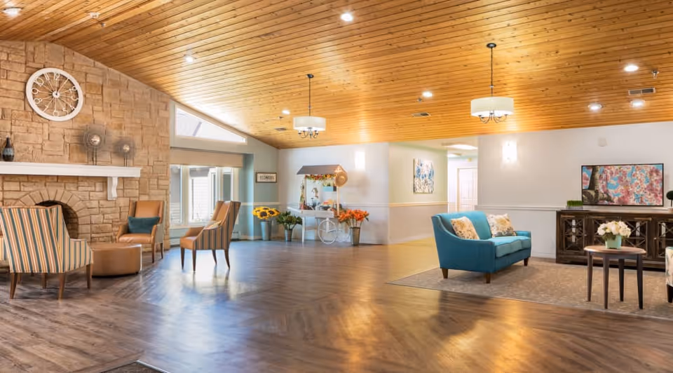 Spacious senior living facility common area with wooden ceiling and recessed lighting. The room features a stone fireplace with a large white clock above it, several chairs including striped and solid-colored ones, a teal loveseat with decorative pillows, a small round table with flowers, and a decorative cart with flowers near the back wall. The floor is a combination of wood and carpet, and the walls are painted light colors with artwork and wall sconces.