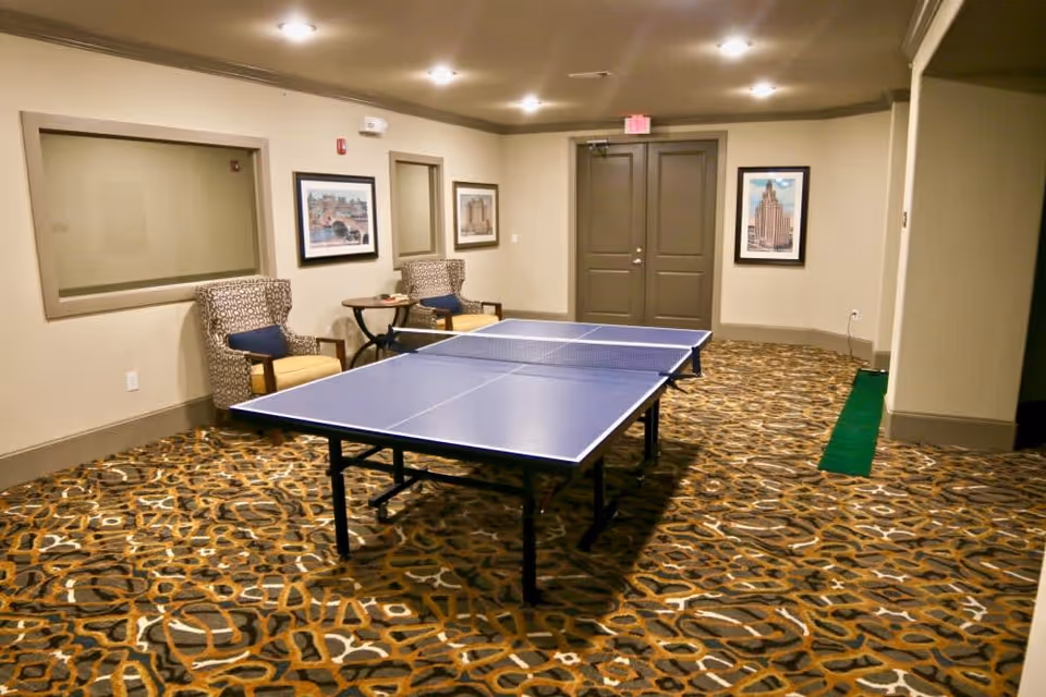 Indoor recreational room with a blue ping pong table in the center, two patterned armchairs with yellow cushions and a small round table against the wall, framed pictures hanging on beige walls, and a patterned carpet covering the floor.