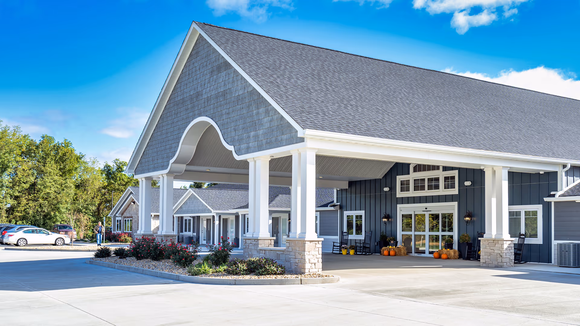 Front exterior view of Villas of Holly Brook Assisted Living & Memory Care: Belleville, showing a large covered entrance with white pillars, stone accents, and a gray roof. There are pumpkins and hay bales near the entrance, some landscaping with bushes and flowers, and a few parked cars in the background under a blue sky with some clouds.