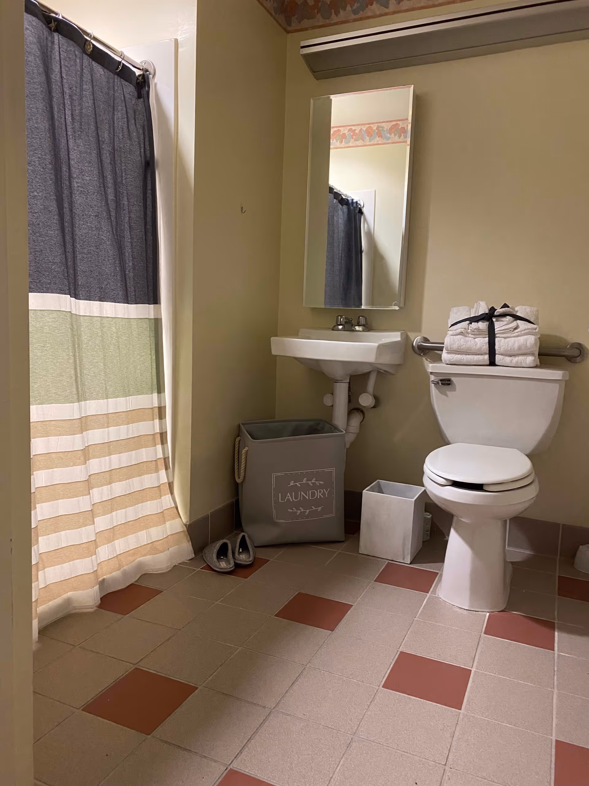 A bathroom with a shower curtain featuring horizontal stripes in dark gray, green, white, and beige. There is a white toilet with folded towels tied with a black ribbon on top, a small white sink with a mirror above it, a gray laundry basket labeled 'LAUNDRY', a small waste bin, and a pair of gray slippers on the tiled floor with a pattern of beige and reddish-brown squares.