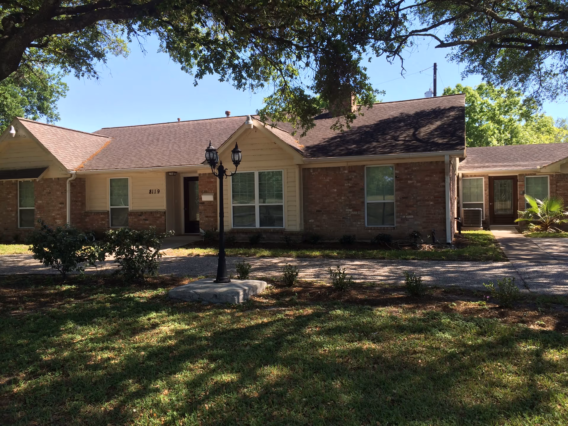 Exterior view of a single-story brick and siding building with multiple windows and doors, surrounded by grass, bushes, and large trees providing shade. A black lamp post is positioned in front of the building.