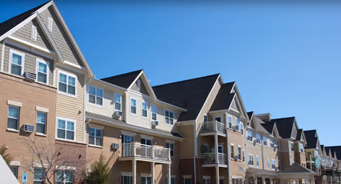 Exterior view of a multi-story senior living facility building with beige and brick facade, multiple windows, balconies, and a clear blue sky in the background.