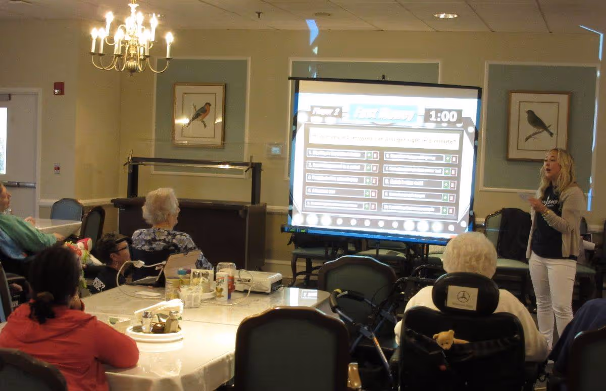 A group of elderly people seated around tables in a common room, watching a woman standing and presenting information displayed on a large screen. The room has framed bird pictures on the walls and a chandelier hanging from the ceiling.