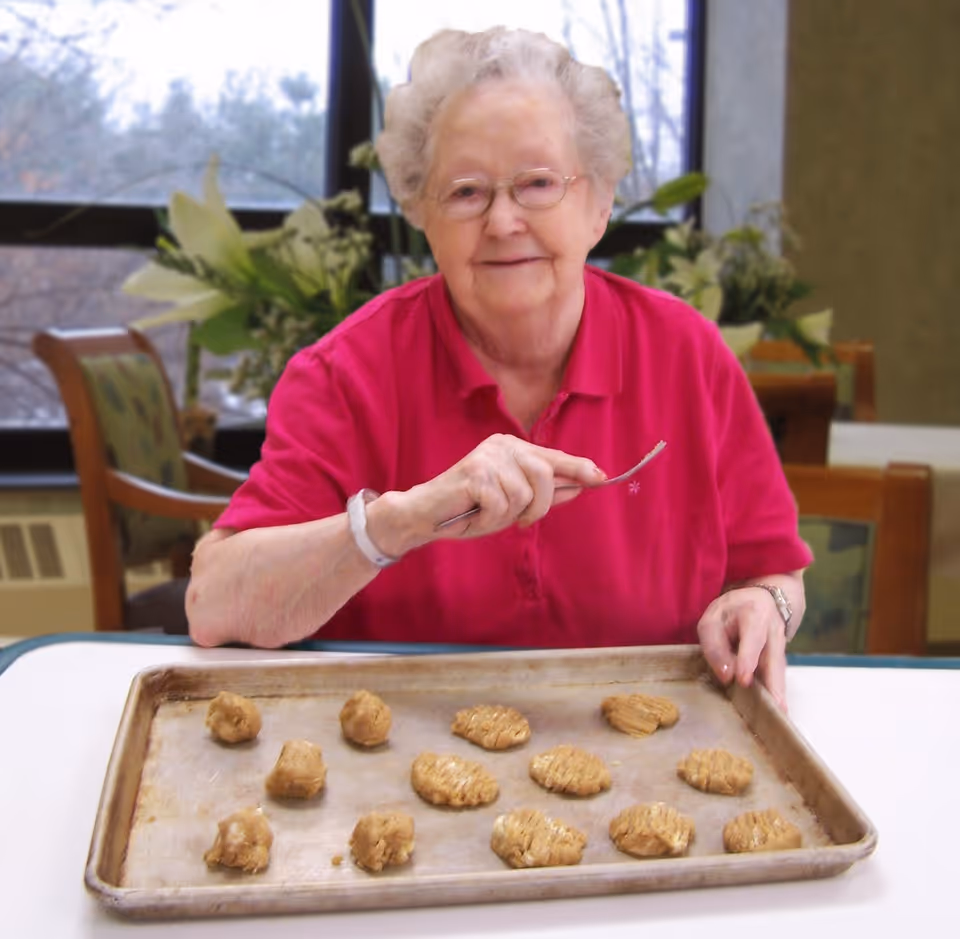 An elderly woman wearing glasses and a bright pink shirt is sitting at a table in a room with large windows and plants in the background. She is holding a fork and smiling at the camera while preparing cookie dough on a baking sheet in front of her.