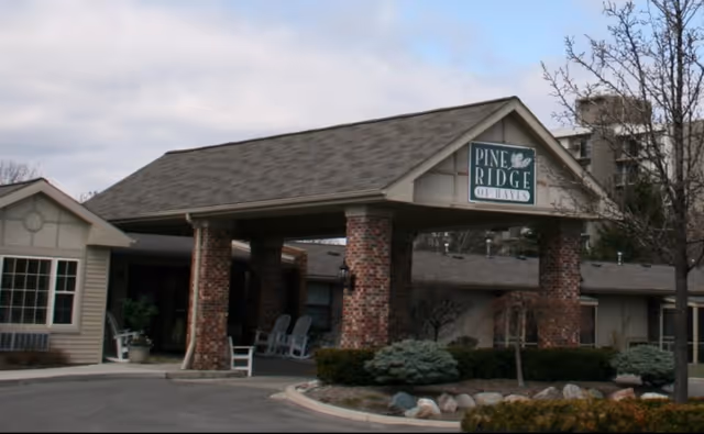 Exterior view of a senior living facility entrance with a covered driveway supported by brick pillars. There are chairs on the porch area and landscaping with bushes and rocks in front. A sign on the covered entrance reads 'Pine Ridge of Dayton'.