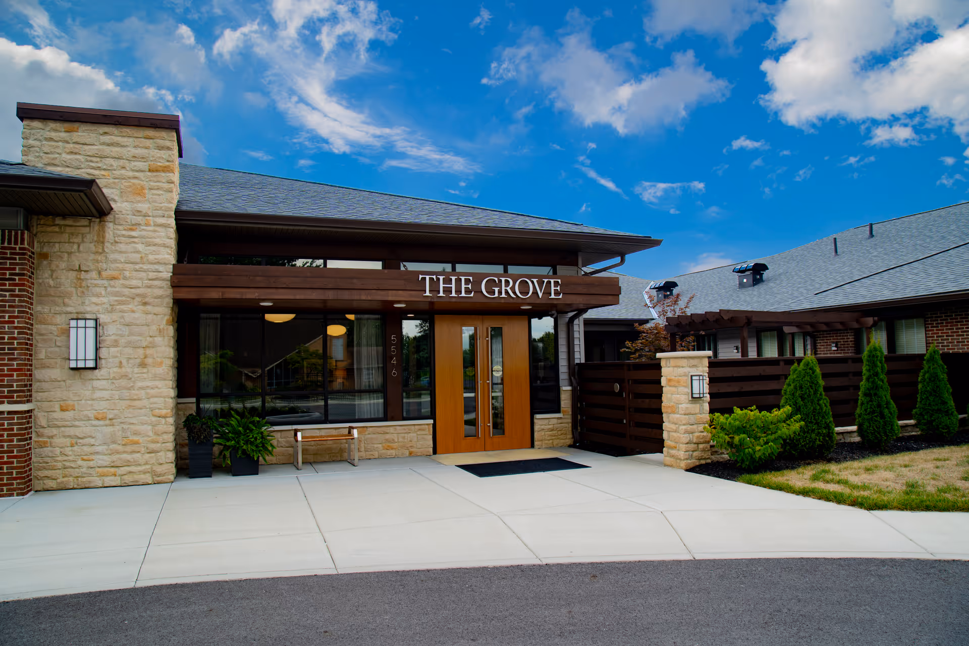 Exterior front entrance of The Grove at Oakleaf Village of Columbus, showing a modern building with stone and brick walls, large glass windows, wooden double doors, and a sign above the entrance that reads 'THE GROVE'. There are plants and shrubs near the entrance and a clear blue sky with some clouds overhead.