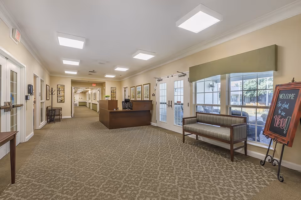 A spacious, well-lit hallway in a senior living facility with a reception desk in the center background. There are framed pictures on the walls, a bench with striped upholstery near large windows on the right, and a chalkboard sign that reads 'WELCOME So glad to see YOU!'. The floor is carpeted with a patterned design, and there are double glass doors leading outside.