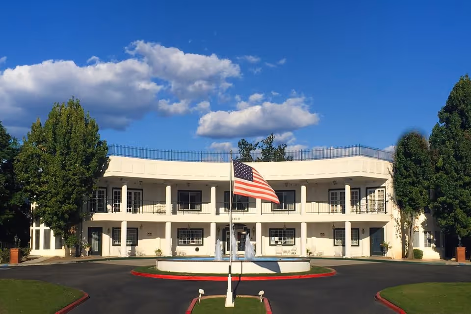 Front exterior view of a two-story white building with a curved facade, featuring balconies with railings on the second floor and several windows and doors on both floors. In front of the building is a circular driveway with a fountain in the center and an American flag on a flagpole. The building is flanked by tall green trees under a blue sky with scattered clouds.