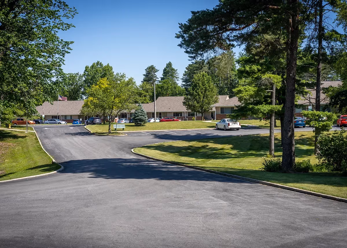 View of a single-story building with a sloped roof surrounded by trees and a well-maintained lawn under a clear blue sky. Several cars are parked in front of the building along a paved driveway.