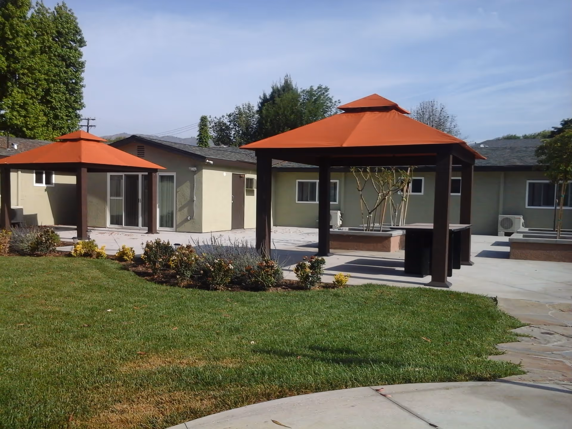 Outdoor area of Glen Park at Glendora featuring two gazebos with orange roofs, surrounded by a well-maintained lawn, flower beds, and a building with beige walls and multiple windows in the background.