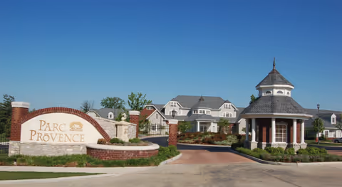 Entrance to Parc Provence senior living facility featuring a large stone and brick sign with the facility name, a circular driveway, landscaped greenery, and a gazebo-style guardhouse with residential buildings in the background under a clear blue sky.