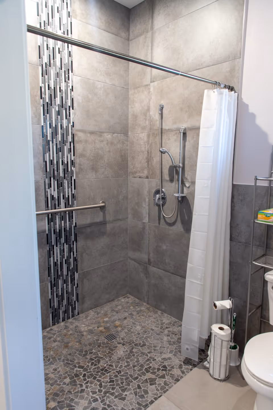 A modern bathroom shower area with gray tiled walls and a mosaic tile strip. The shower floor has pebble-style tiles and a metal grab bar is installed on the wall. A white shower curtain is partially drawn, and a handheld showerhead is mounted on the wall. To the right, part of a toilet and a metal shelving unit with bathroom supplies are visible.