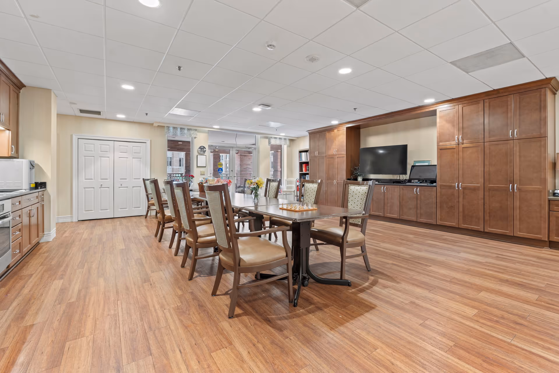 A spacious room with wooden flooring and a long table surrounded by chairs. The room features built-in wooden cabinets along one wall, a flat-screen TV, and a microwave on the left side. Large windows and glass doors at the far end let in natural light.