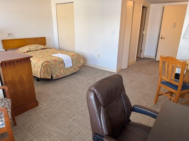 A spacious assisted living bedroom with a single bed covered in a floral bedspread, a wooden dresser, a brown leather office chair, a wooden dining chair, and a table. The room has beige carpet and white walls, with a closet and a hallway visible in the background.