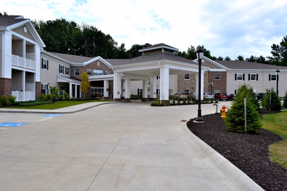 Exterior view of a senior living facility named Danbury Mentor featuring a large covered entrance with white pillars, a driveway, landscaped areas with small trees and shrubs, parking spaces including handicapped spots, and a multi-story building with beige siding and brick accents under a partly cloudy sky.