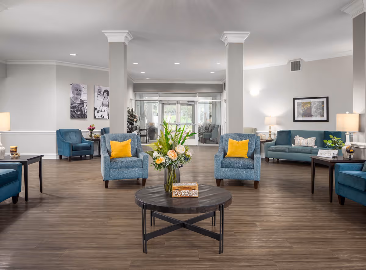 A spacious and well-lit senior living facility common area with wooden flooring, blue upholstered chairs and sofas, yellow accent pillows, a round coffee table with a flower arrangement, side tables with lamps, and black and white portraits on the wall. The room has large glass doors at the far end and neutral-colored walls.