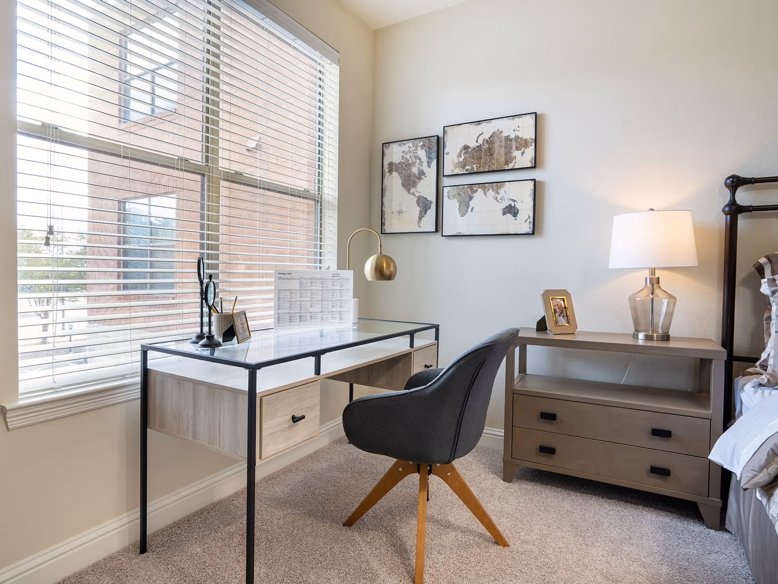 A bright room with a large window covered by white blinds. In front of the window is a modern desk with a glass top and wooden drawers, accompanied by a dark gray chair with wooden legs. On the desk are decorative items including a small sculpture, a pencil holder, and a calendar. On the adjacent wall are three framed world map artworks. Next to the desk is a wooden nightstand with two drawers, a photo frame, and a table lamp with a white shade. Part of a bed with pillows is visible on the right side.