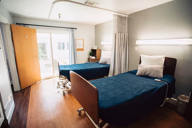 A bright room with two hospital-style beds covered in dark blue bedding, each with white pillows. The room has wooden flooring, a partially open wooden door leading outside, a window with light curtains, a small wooden nightstand with a plant and a phone, and a privacy curtain between the beds.