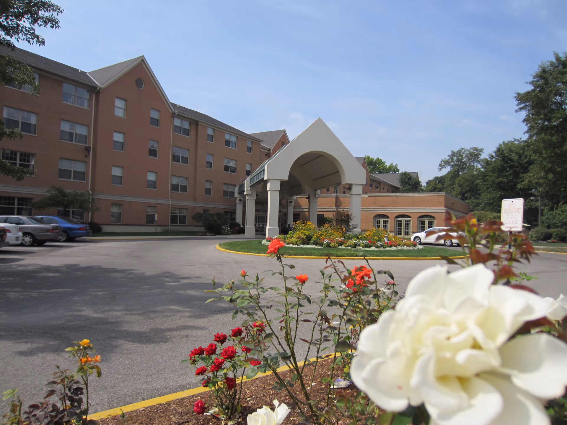 Exterior view of a multi-story brick building with a covered entrance and a circular driveway. There are colorful flowers in the foreground and several parked cars near the building. Trees and a clear sky are visible in the background.