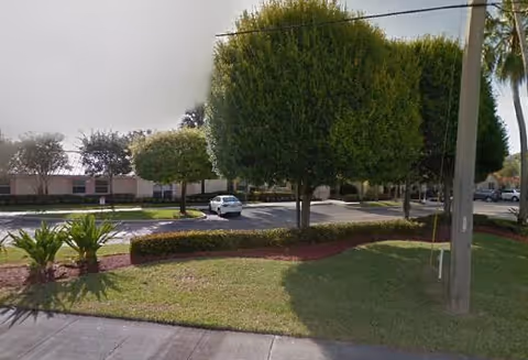 A parking lot area with neatly trimmed trees and landscaped grass patches in front of a single-story building, under a partly cloudy sky.
