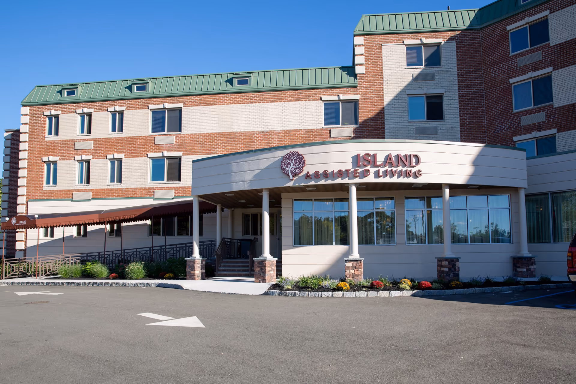 Exterior front view of a multi-story brick building with a green roof and a covered entrance. The entrance has a sign that reads 'Island Assisted Living' with a tree logo. There are windows on all floors and a paved driveway with a white directional arrow in front.