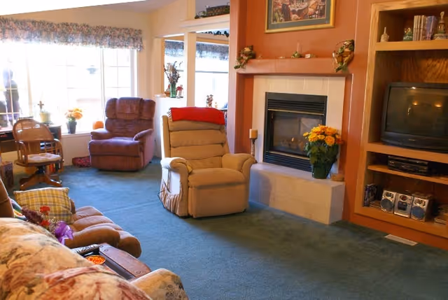 A cozy living room in a senior care facility featuring a beige recliner chair with a red blanket draped over it, a brown armchair, a floral patterned sofa, a fireplace with a vase of yellow flowers on the hearth, built-in wooden shelves holding a TV and stereo system, and large windows with floral valances letting in natural light.