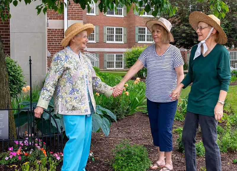 Three elderly women wearing hats stand outside in a garden area holding hands and smiling. They are surrounded by green plants and flowers, with a brick building visible in the background.