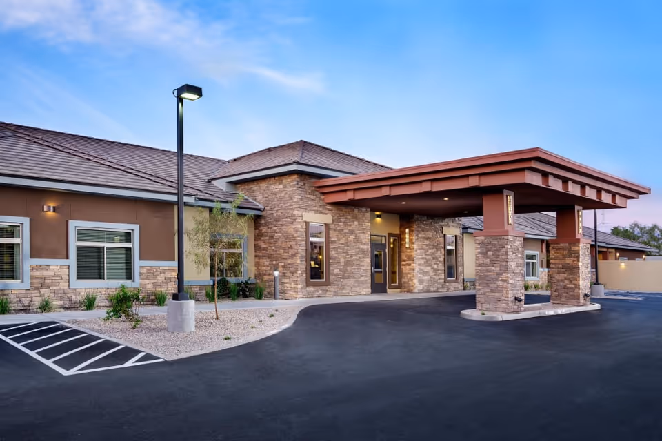 Exterior view of a single-story building with stone and stucco walls, a covered entrance supported by stone pillars, a parking area with marked spaces, and a street lamp, under a clear blue sky at dusk.