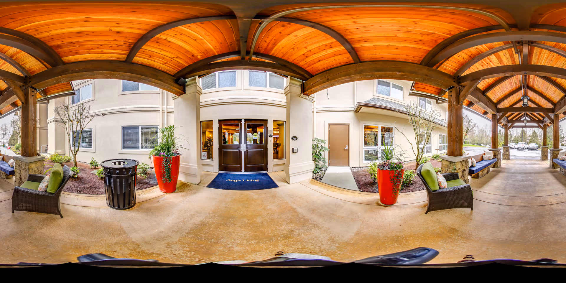 Covered outdoor entrance area of a senior living facility with wooden beams and ceiling, stone pillars, wicker chairs with green cushions, large red planters with greenery, and double glass doors leading inside. The building exterior is beige with several windows, and there is a blue mat with the text 'Aegis Living' in front of the doors.