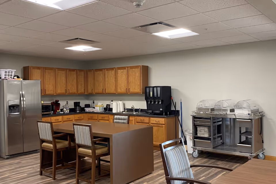 A kitchen area with wooden cabinets, a stainless steel refrigerator, a countertop with various appliances including a coffee maker and a water dispenser. There is a brown island table with four chairs around it and a metal serving cart with covered trays on the right side. The floor is wood-patterned and the ceiling has recessed lighting.
