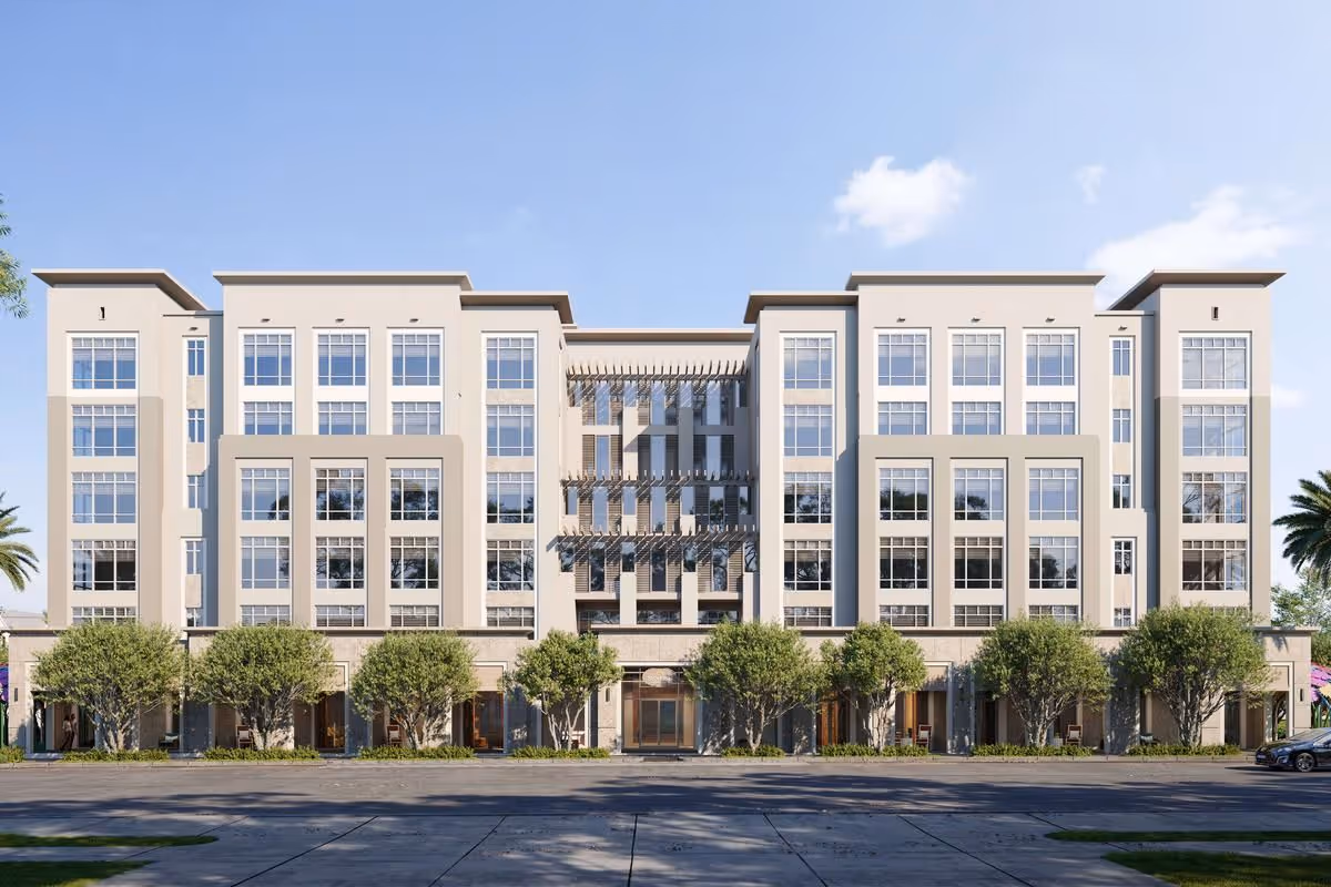Front exterior view of a modern multi-story senior living facility building with large windows, trees lining the entrance, and a clear blue sky.