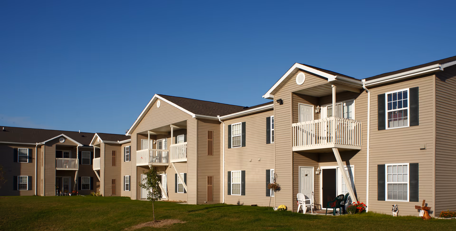 Exterior view of a two-story beige apartment building with white balconies and multiple windows under a clear blue sky. There is a green lawn in front of the building with a small tree and some outdoor chairs on the ground floor patios.