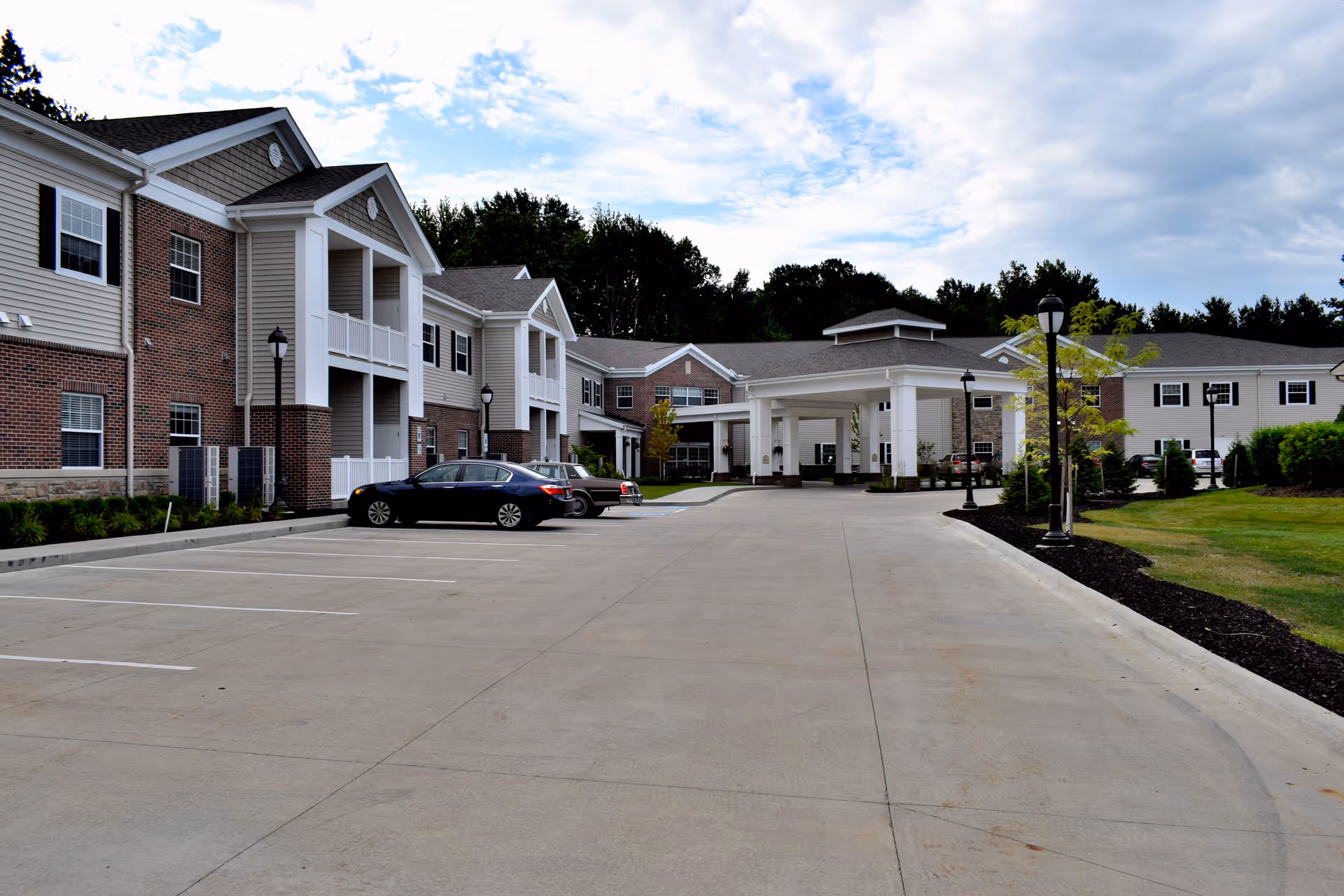 Exterior view of a senior living facility named Danbury Mentor showing a large driveway and parking area with a few cars parked. The building is two stories with a combination of brick and siding, featuring balconies and a covered entrance. There are lamp posts and landscaped greenery around the driveway.