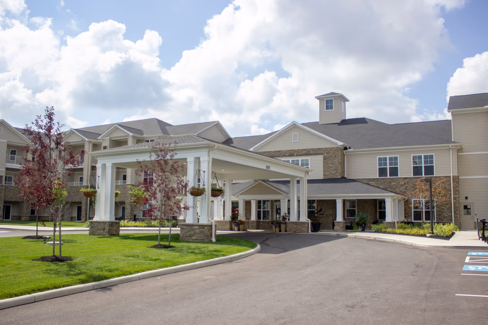 Exterior view of StoryPoint Grove City senior living facility showing a large building with beige siding and stone accents, a covered entrance with white columns, a driveway, green lawn, and small trees under a partly cloudy sky.
