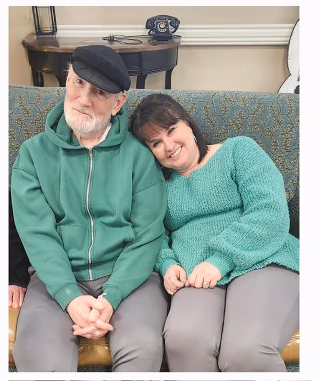 An elderly man wearing a black cap and green zip-up hoodie sits on a patterned couch next to a woman in a green sweater who is leaning her head on his shoulder, both smiling. Behind them is a wooden table with a vintage black rotary phone on it.