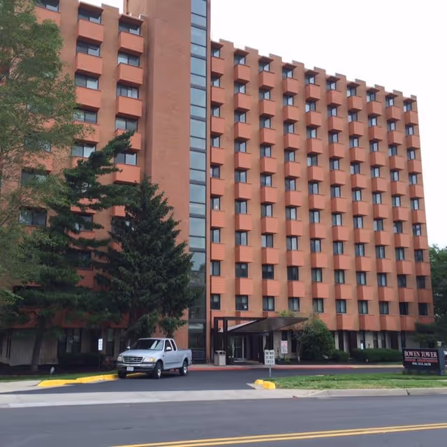 Exterior view of Bowen Tower Senior Independent Living Apartments, a multi-story brick building with rows of windows and small balconies. There are trees and a parked silver pickup truck near the entrance, with a sign displaying the facility name on the lawn.